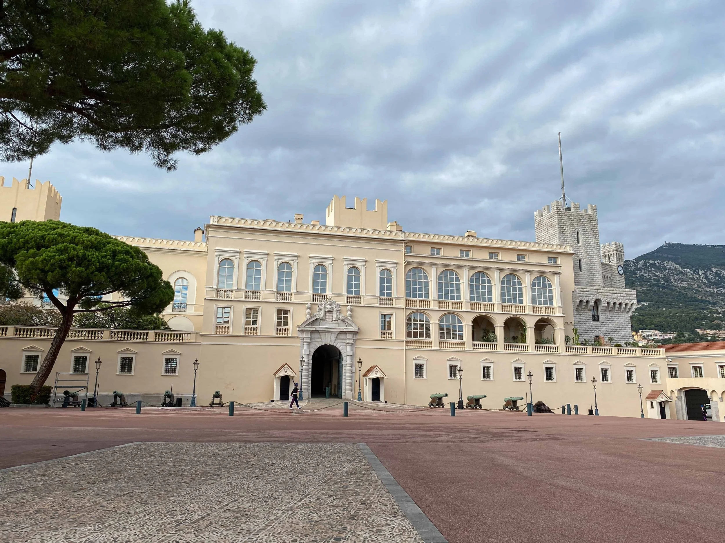 The Prince's Palace of Monaco on Place du Palais — a cream-colored Baroque facade with medieval towers, a lone Palace Guard at the entrance, and dramatic storm clouds over the Rock