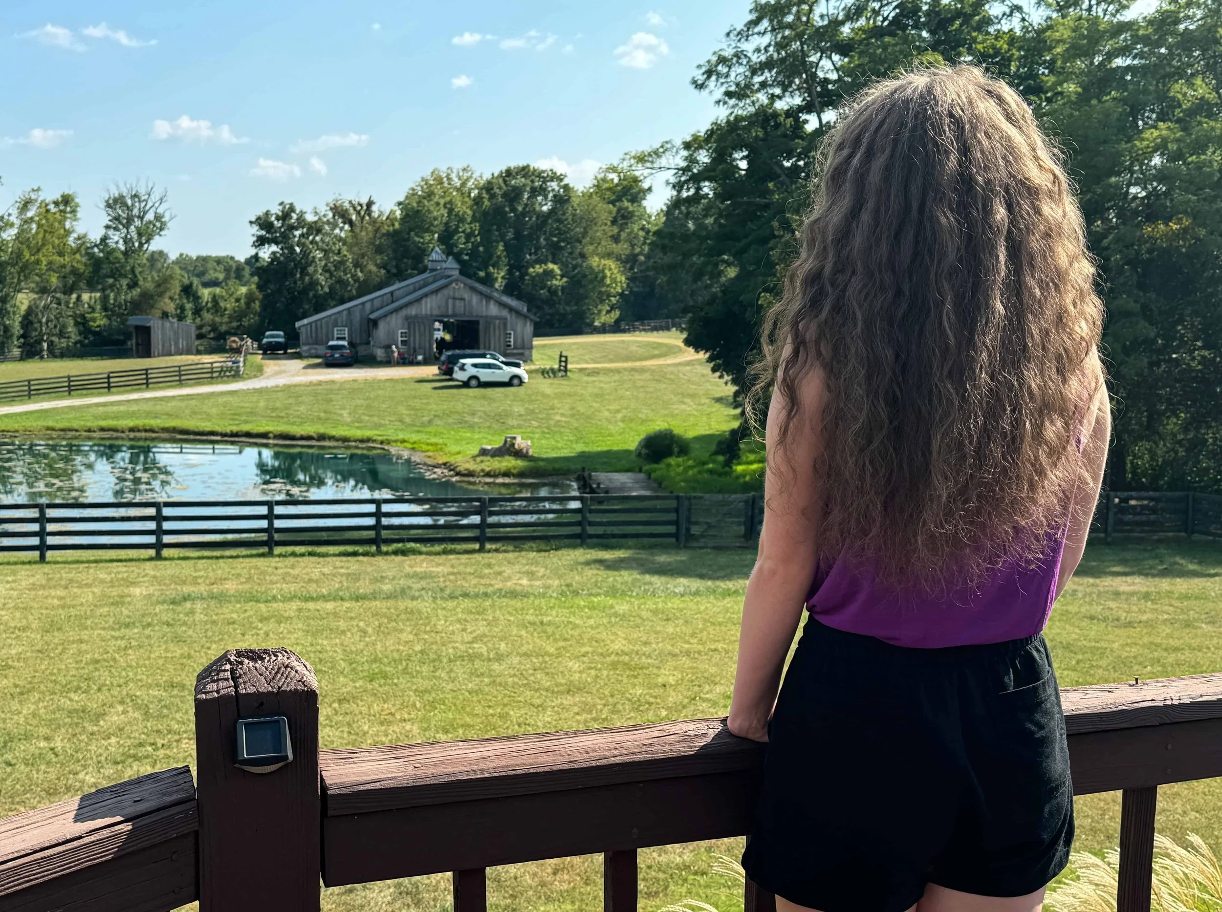 A girl with long curly hair wearing a purple top and black shorts standing on a wooden deck, looking out over a green field with a pond, a fence, trees, and a rustic barn in the distance on a sunny day.