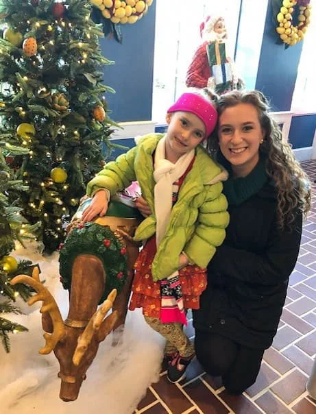 Two family members pose with a reindeer statue beside a decorated Christmas tree in a restaurant lobby.