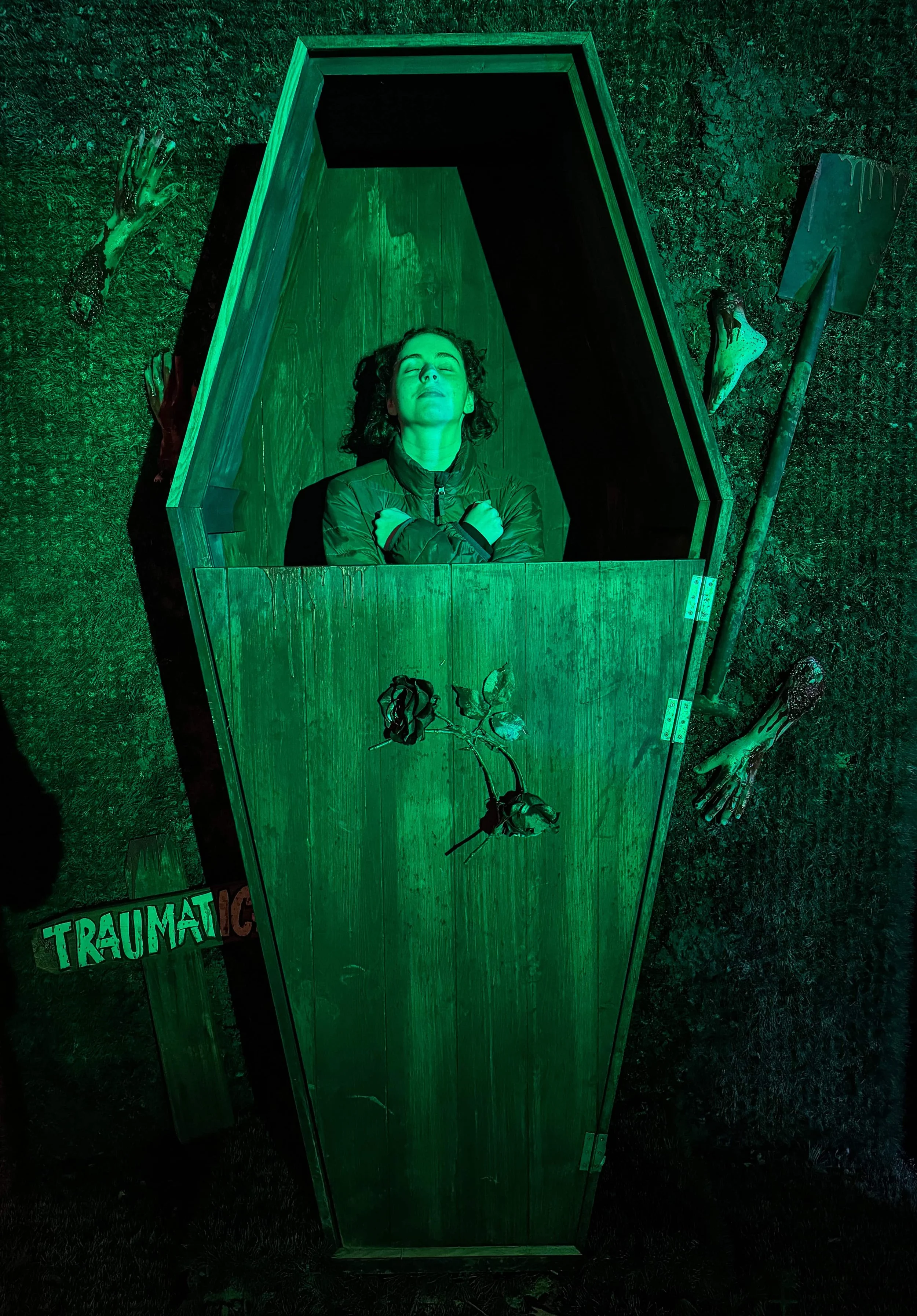 A family member lying inside a green-lit coffin prop, a photo-op display at Traumatica.
