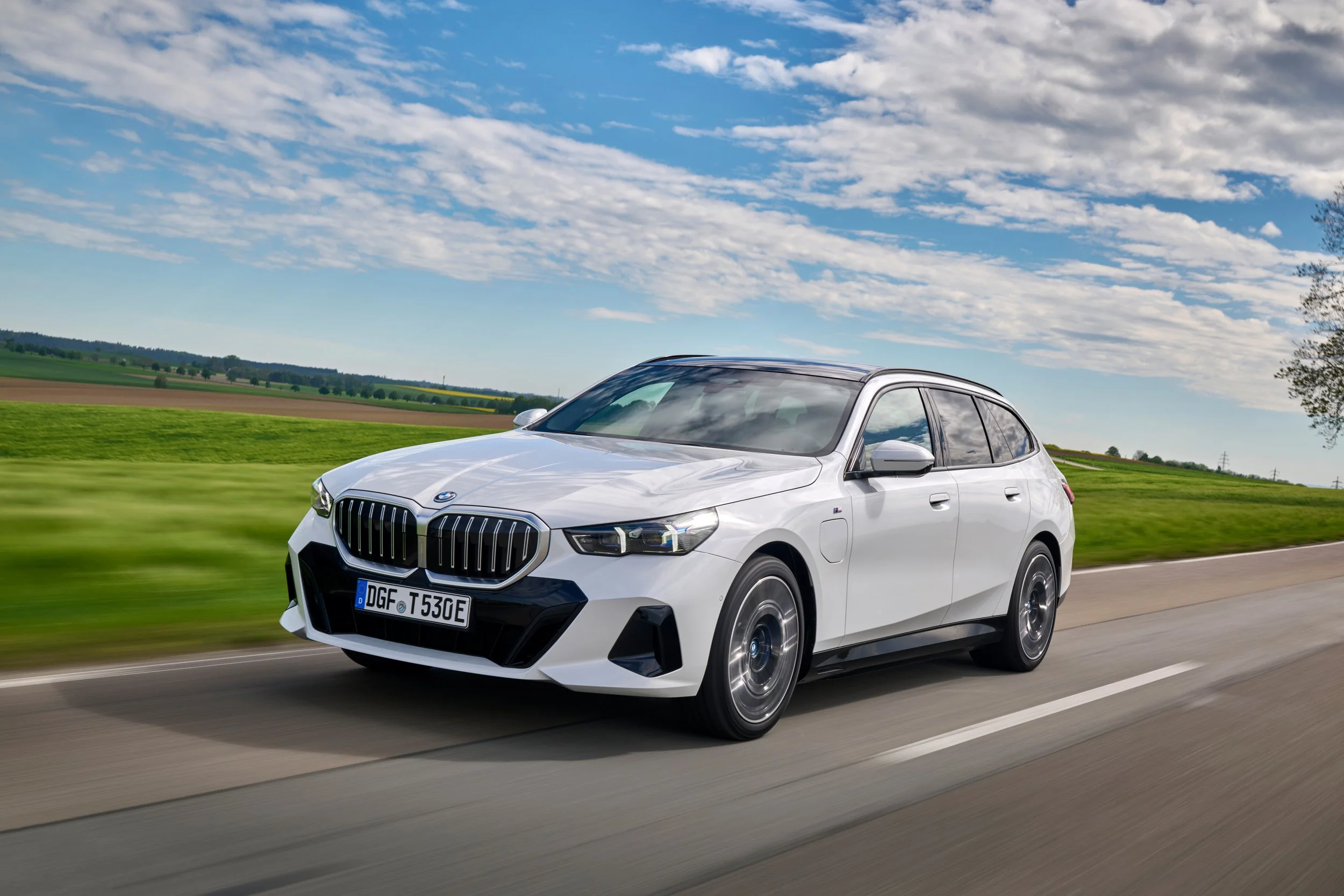 A white BMW SUV driving on a rural road with green fields and a partly cloudy sky in the background.