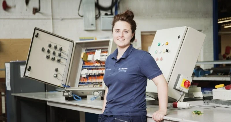 Woman in navy blue polo shirt standing in front of electrical control panels in a workshop or industrial setting.