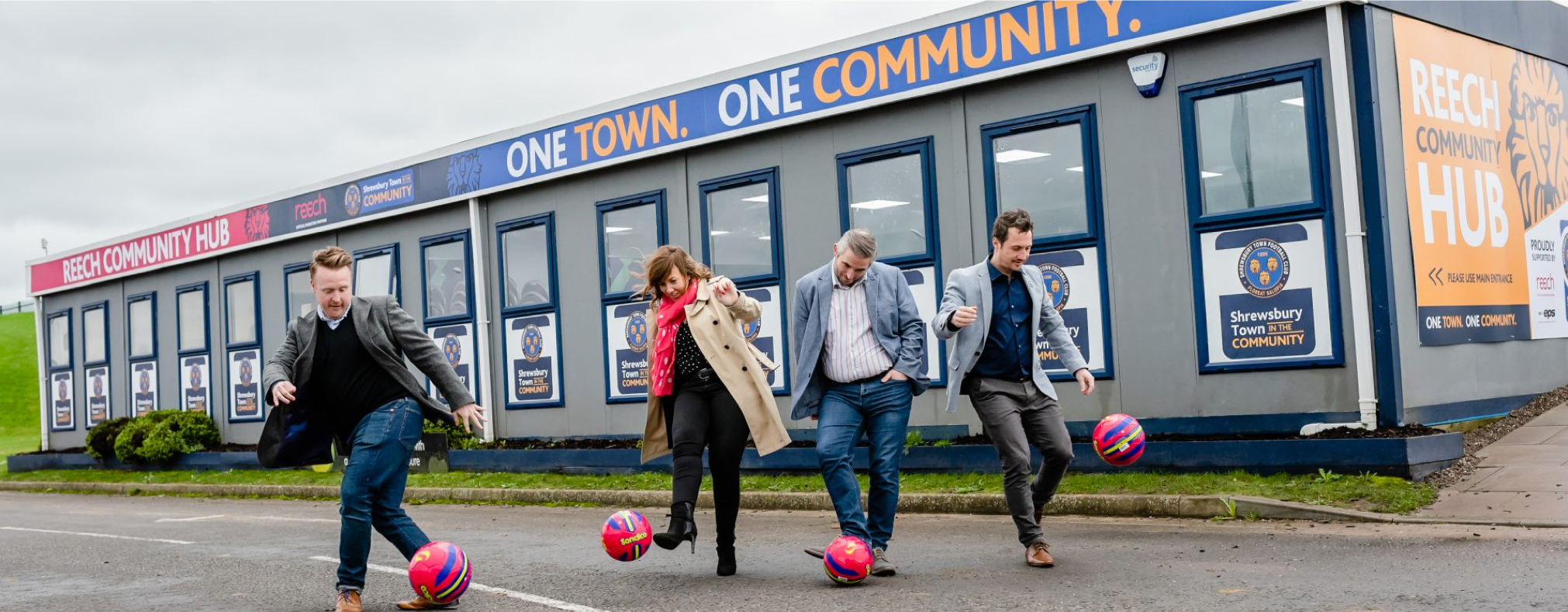Four people playing with colorful soccer balls in front of a community hub building, with banners reading 'Reech Community Hub' and 'One Town. One Community.'