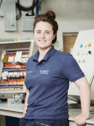Woman in navy blue polo shirt standing in front of electrical control panels in a workshop or industrial setting.
