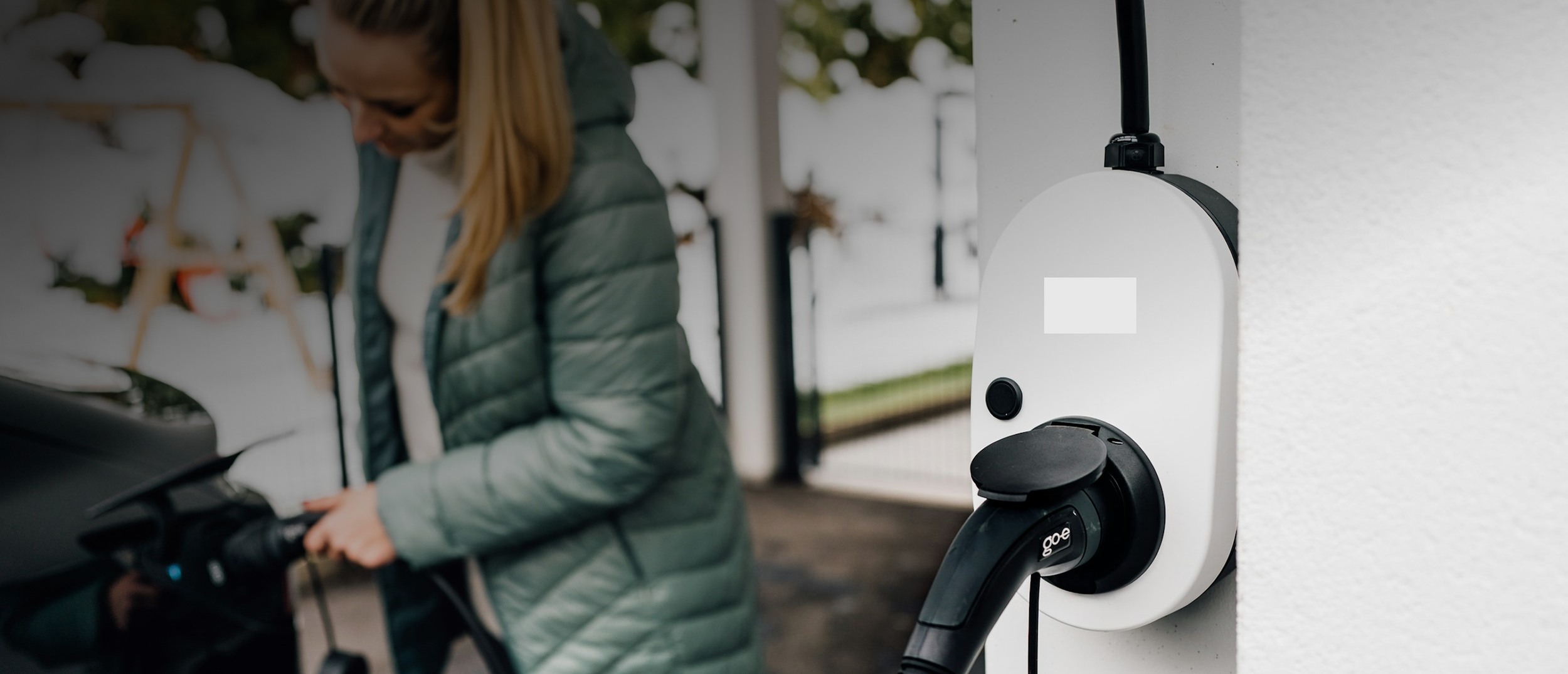 Woman in a gray jacket charging an electric vehicle at an outdoor charging station.
