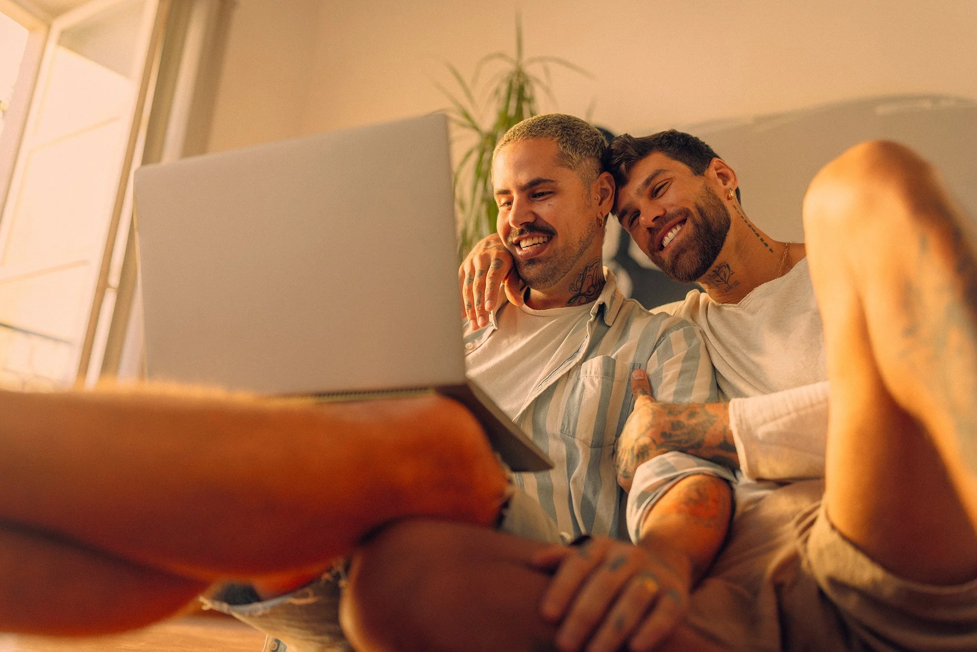 Two young men with tattoos, smiling and enjoying each other's company while watching something on a laptop in a cozy room.