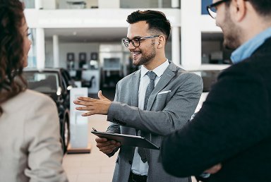 A man in a gray suit and glasses smiling and talking with two people in a car dealership.