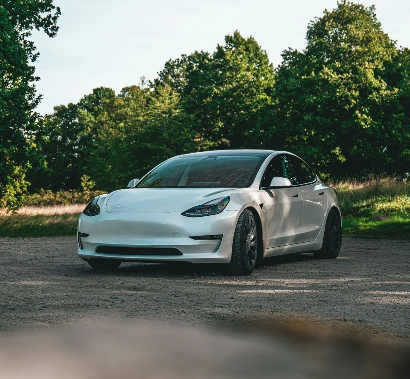 A white Tesla Model 3 car parked on a dirt road with green trees in the background.