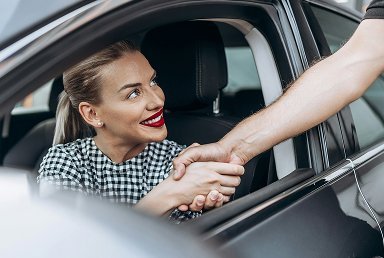 A woman with blonde hair and red lipstick smiling and shaking hands with a person outside a car through the open window.