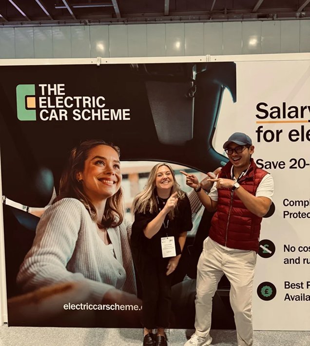 Three people in front of a large banner for the Electric Car Scheme, smiling and pointing at each other, inside a venue or trade show setting.