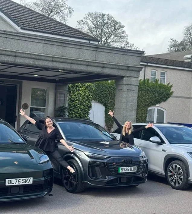 Two women standing beside luxury cars with their arms raised in a cheerful manner in front of a residence with a garage, a stone facade, and greenery.