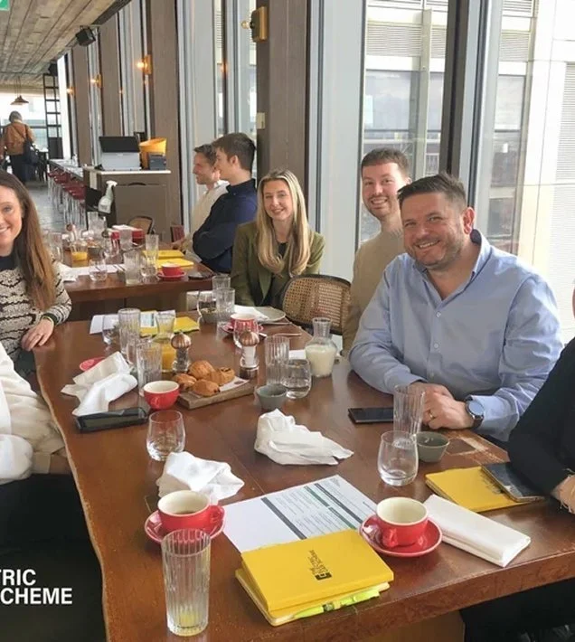 Group of people sitting around a long dining table during breakfast, with food, drinks, and tableware, in a modern restaurant with large windows