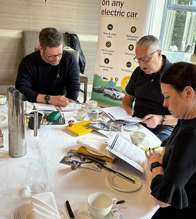 Three people sitting at a conference table, reviewing documents and taking notes in a meeting. The table has cups, glasses, folders, and writing materials, with a promotional banner about electric cars in the background.