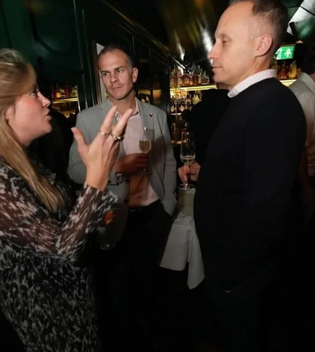 Three people engaged in conversation at a social event, holding drinks, in a dimly lit venue.