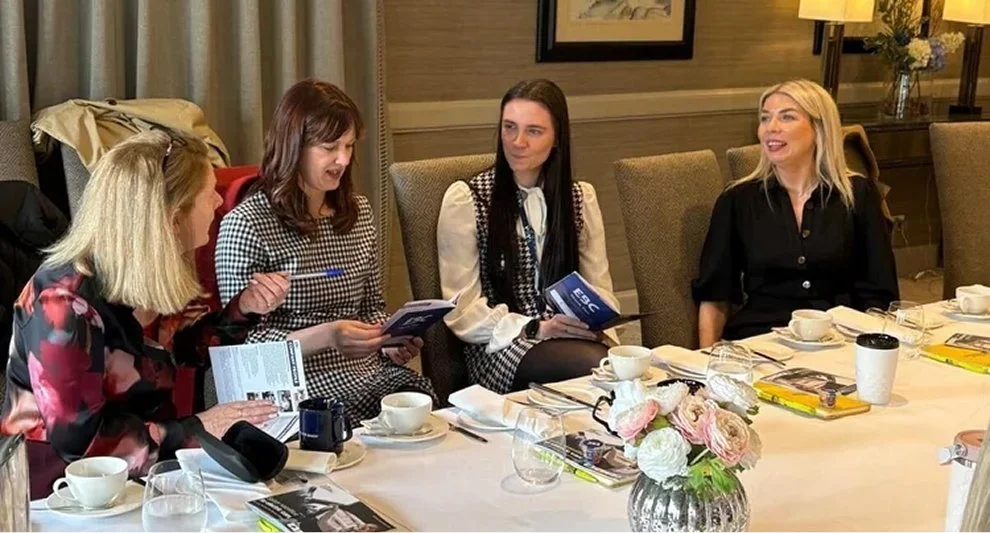 Five women sitting at a dinner table with cups, glasses, books, and a floral centerpiece, engaging in conversation in a well-lit room.