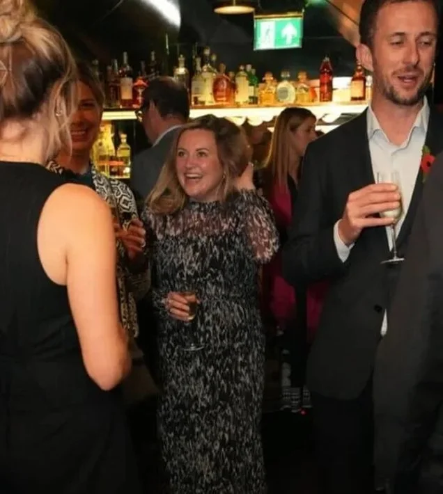 Group of people socializing at a bar, with bottles of alcohol on the shelf in the background, some holding drinks, in a lively and festive atmosphere.