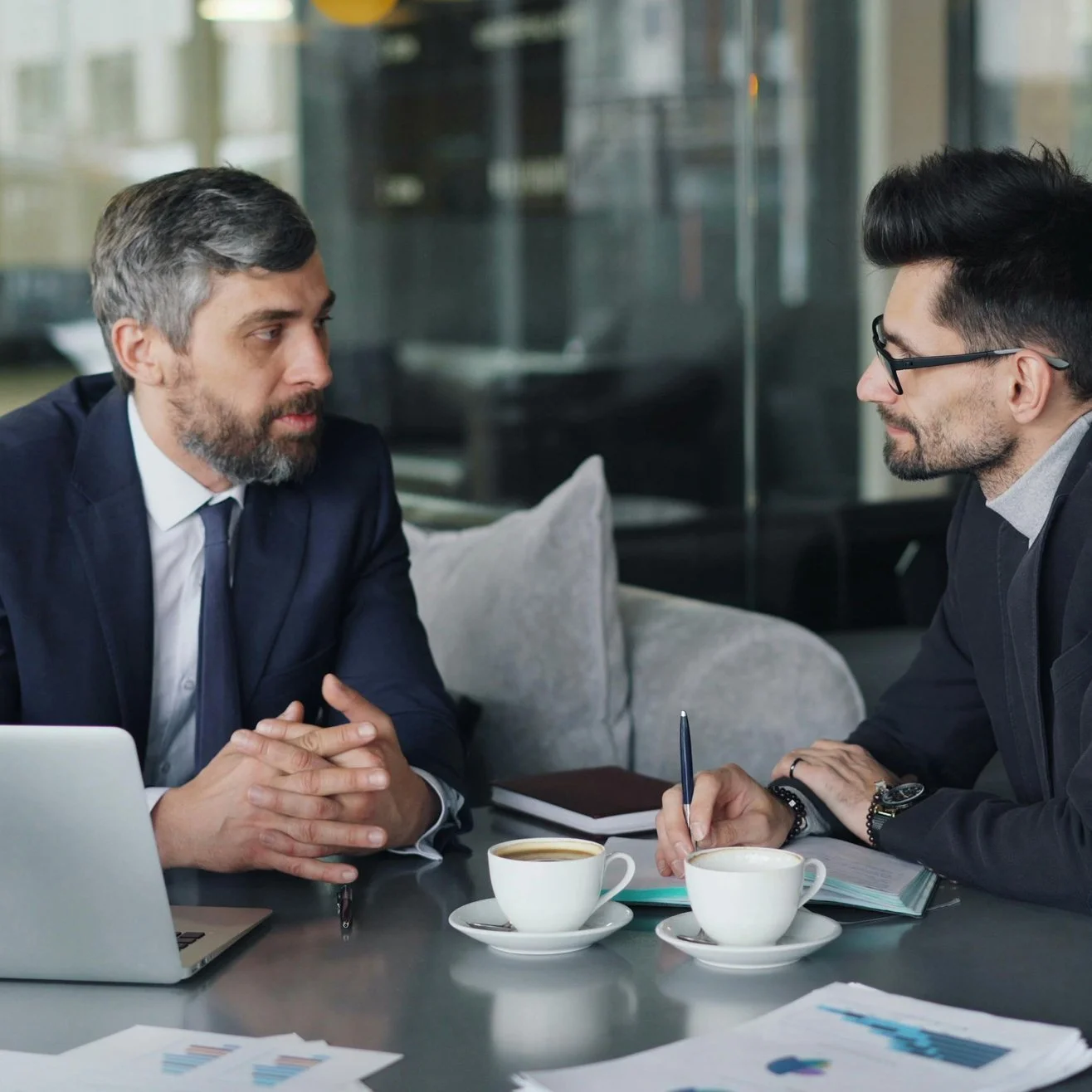 Two men in business suits having a serious discussion at a meeting table with coffee cups, laptop, notebooks, and charts.