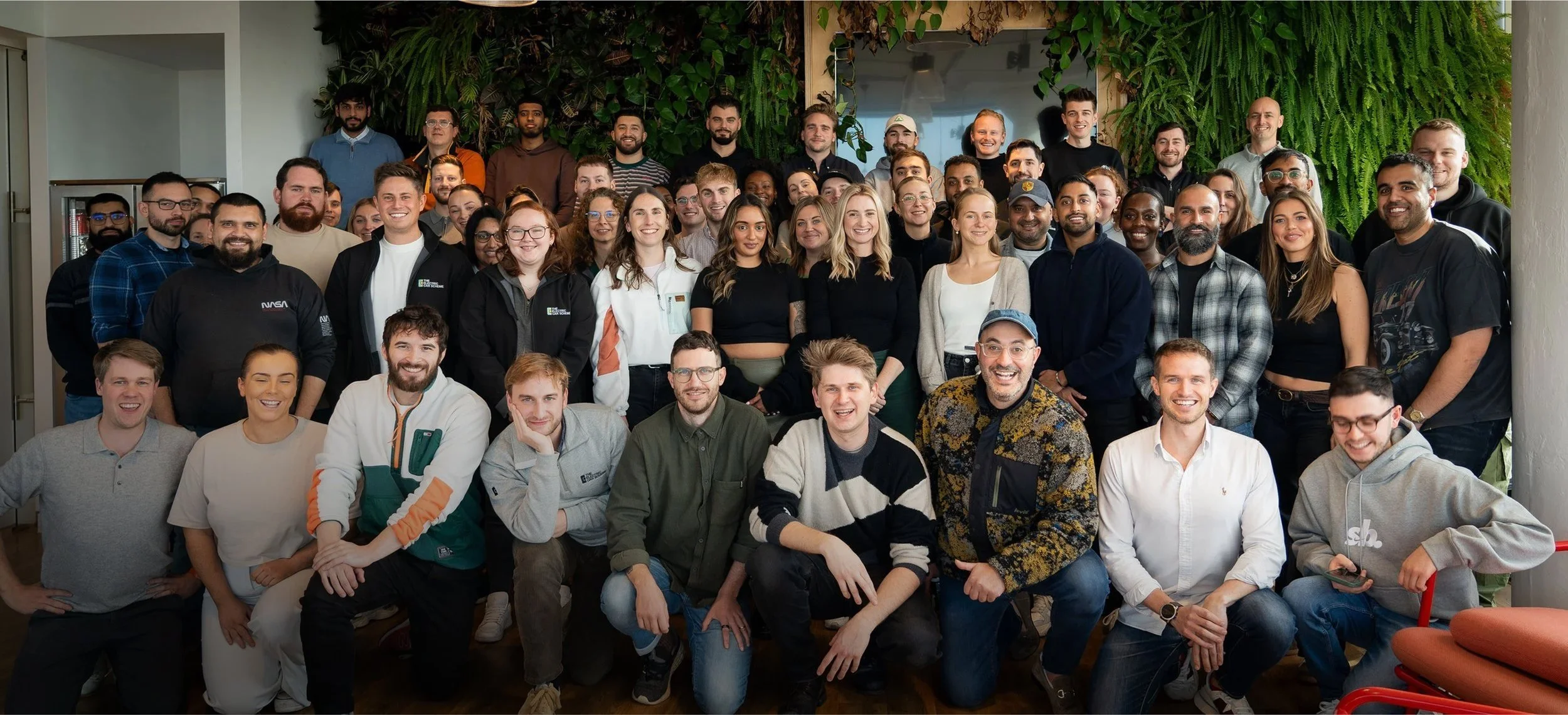 Group photo of diverse people smiling and posing together indoors, with green plants in the background.