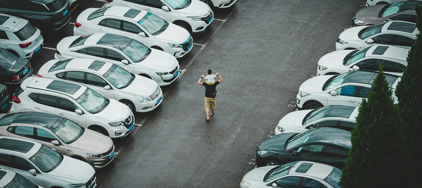A man carrying a young boy on his shoulders walking through a parking lot filled with mostly white and gray cars.