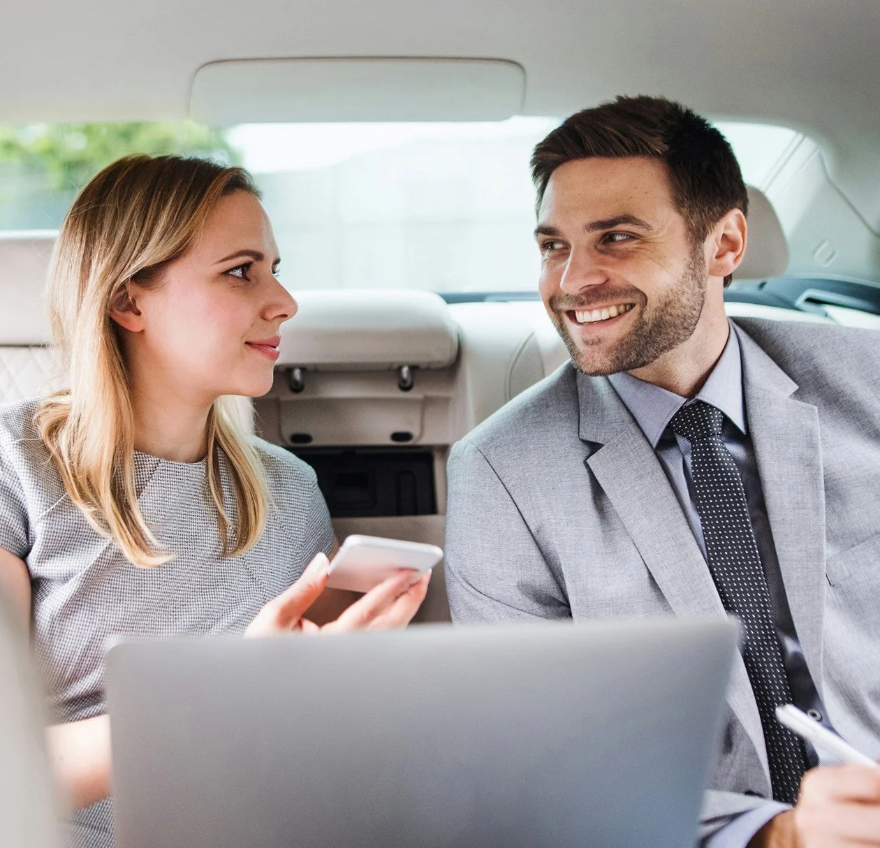 A woman and a man talking and smiling in the backseat of a car, with the woman holding a smartphone and a laptop in front of them.