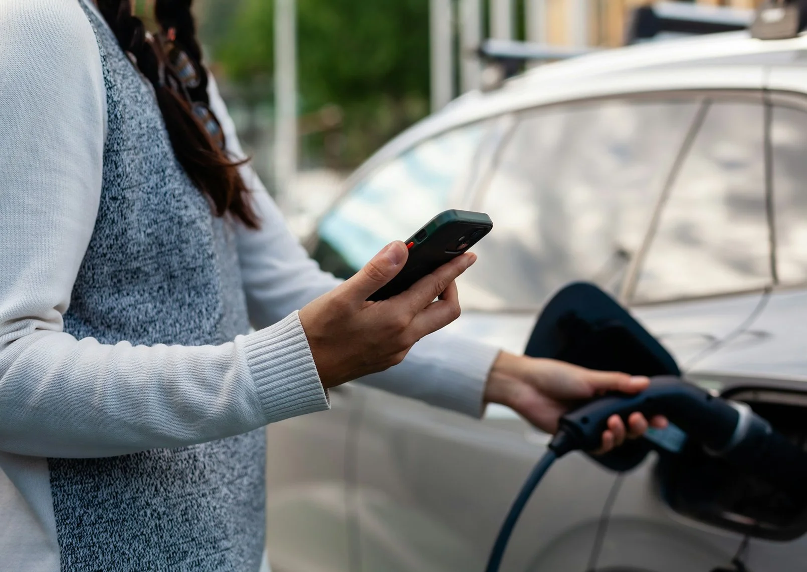 A person standing next to an electric vehicle charging it, holding a smartphone in one hand and the charging cable in the other.
