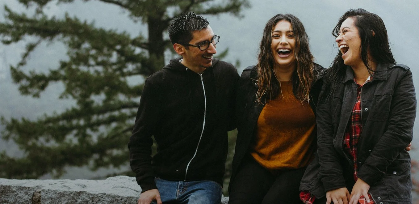 Three friends smiling and laughing outdoors in front of a tree and cloudy sky, enjoying each other's company.