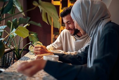A man and a woman sitting at a table in a cozy indoor space, smiling and looking at a smartphone together, surrounded by green plants.