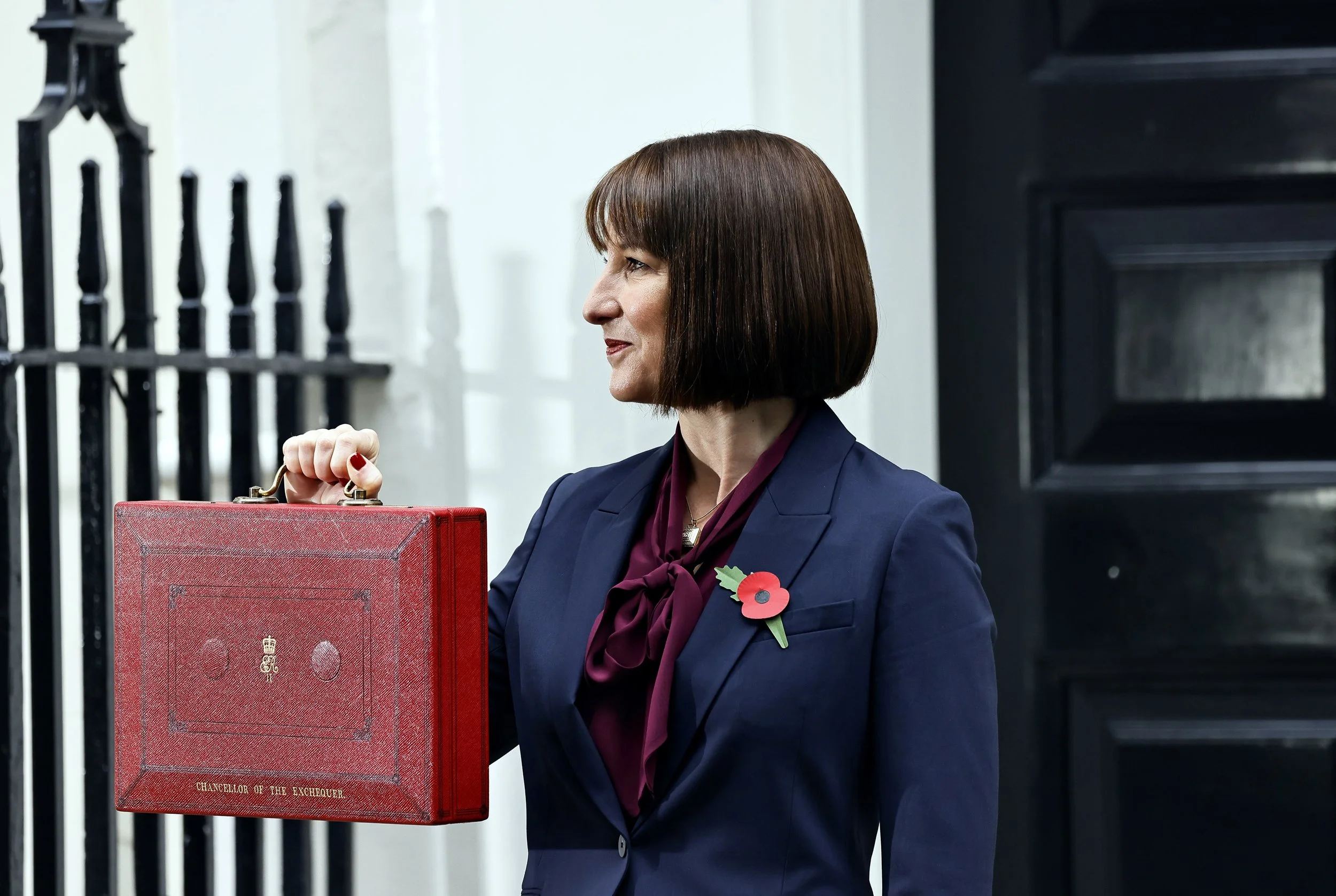 A woman with shoulder-length brown hair holding a red briefcase labeled 'Chancellor of the Exchequer' outside a building with black door and white wall, wearing a navy blazer with a poppy pin and a burgundy scarf.