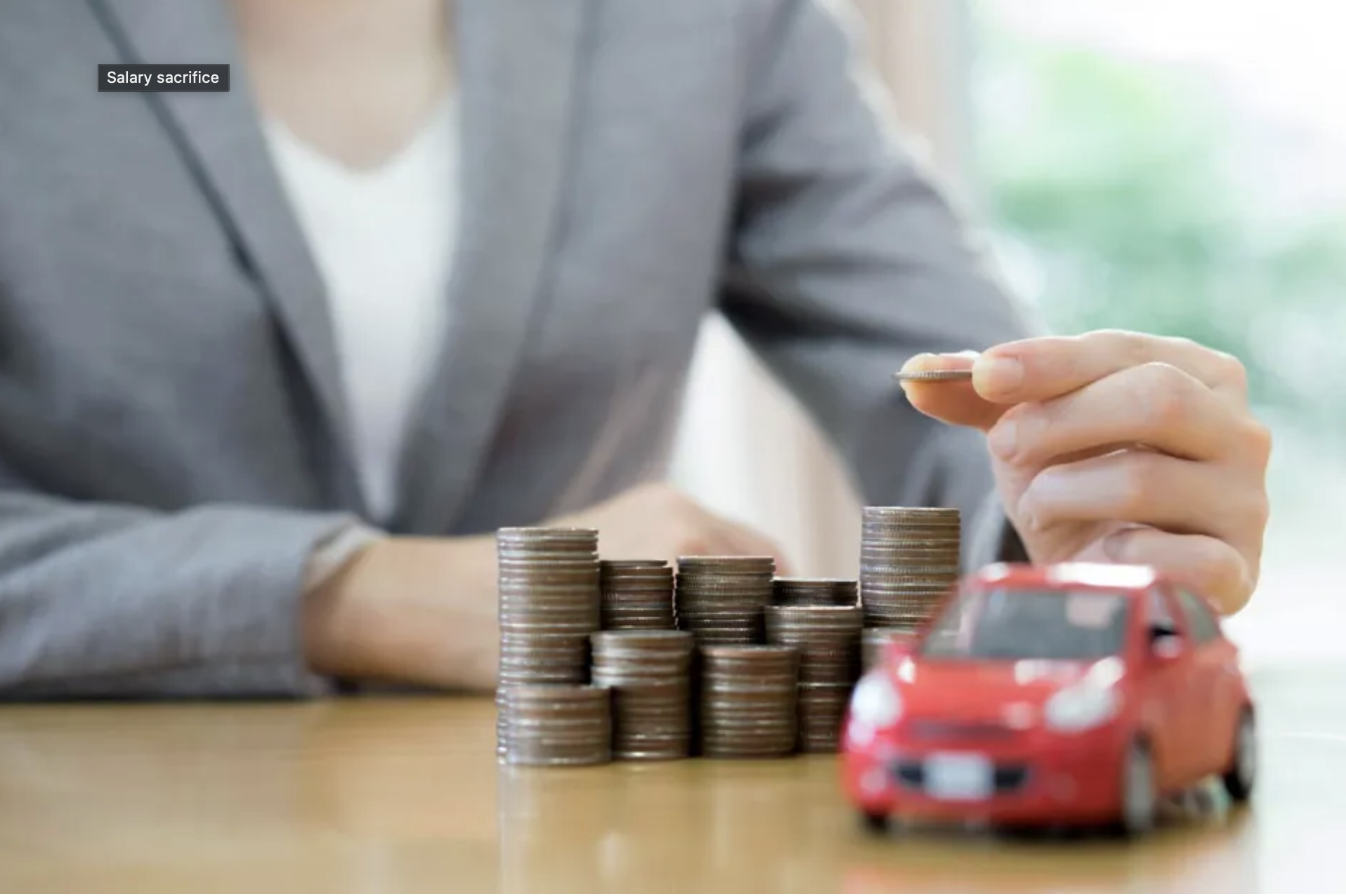 A person in a grey blazer stacking coins on a table, with a small red toy car in front of the coins. The person is holding a coin in their right hand, and there are multiple stacks of coins of varying heights. A label on the photo reads "Salary sacrifice."