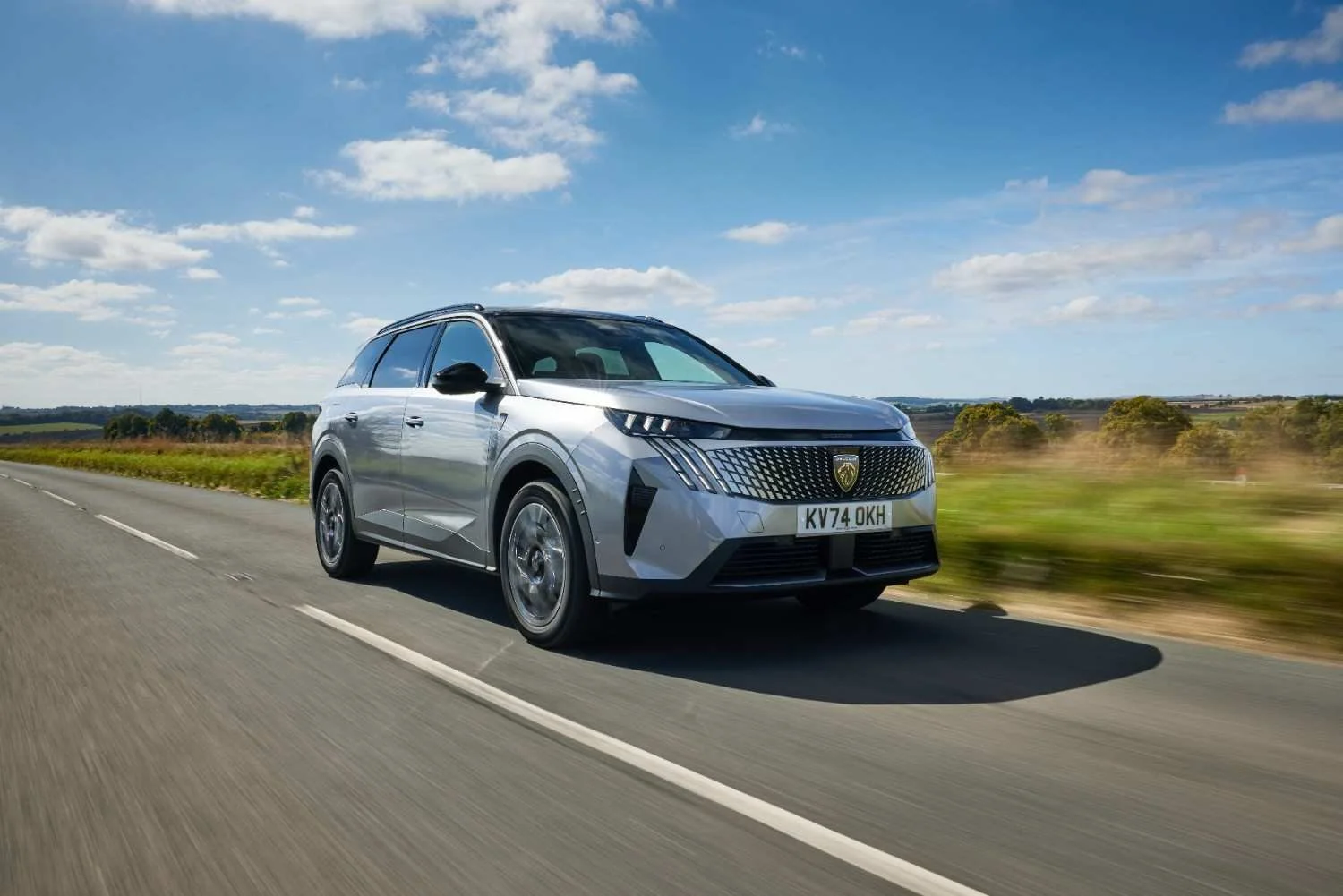 A silver SUV driving on a two-lane highway through a rural landscape under a partly cloudy sky.