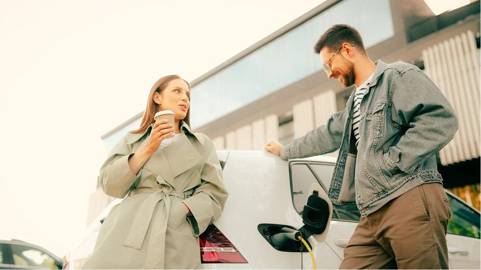 A woman drinking coffee and a man smiling next to an electric car plugged into a charging station outside a modern building.