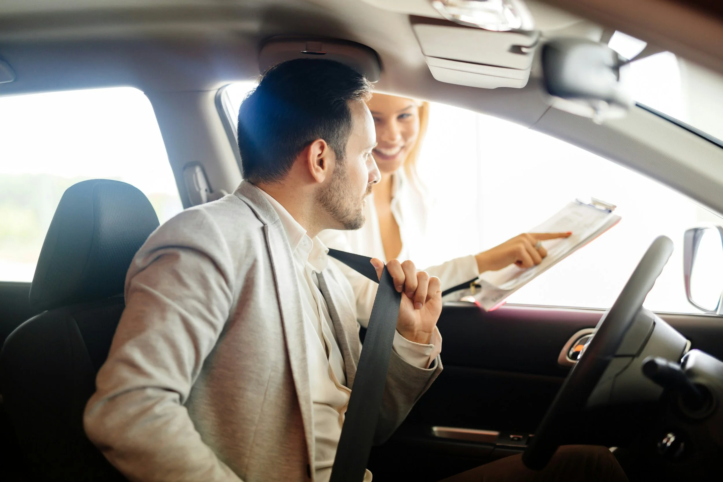 A man is sitting in the driver's seat of a car, and a woman is standing outside the car, leaning in and pointing at something on a clipboard or tablet. The woman is smiling, and both are engaged in a conversation inside the vehicle.