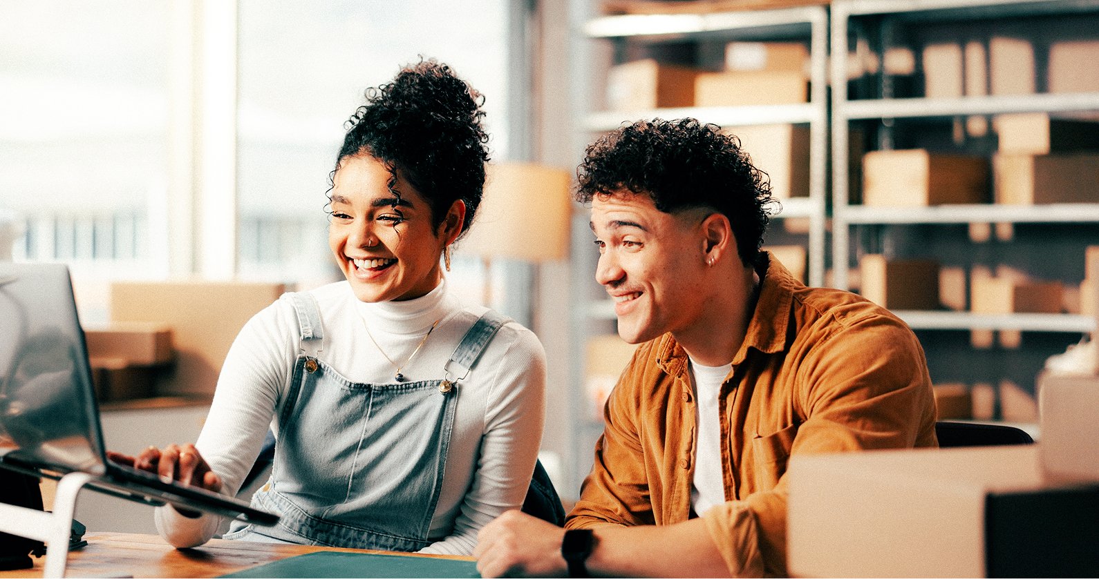 Two young adults, a woman and a man, smiling and looking at a laptop in a brightly lit office or workspace.