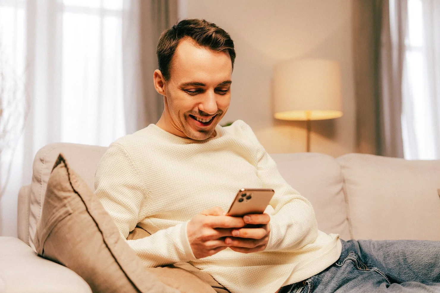 A young man with short dark hair and a light complexion, wearing a cream sweater, sitting on a beige sofa in a cozy living room, looking at his smartphone and smiling.