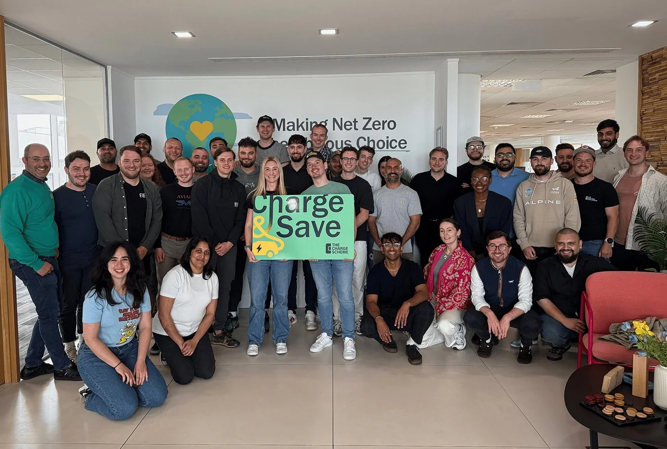 Group of smiling people gathered in an office, holding a sign that reads "Charge & Save," with a globe and a yellow heart logo on the wall behind them. The wall also displays the text "Making Net Zero Conscious Choice."
