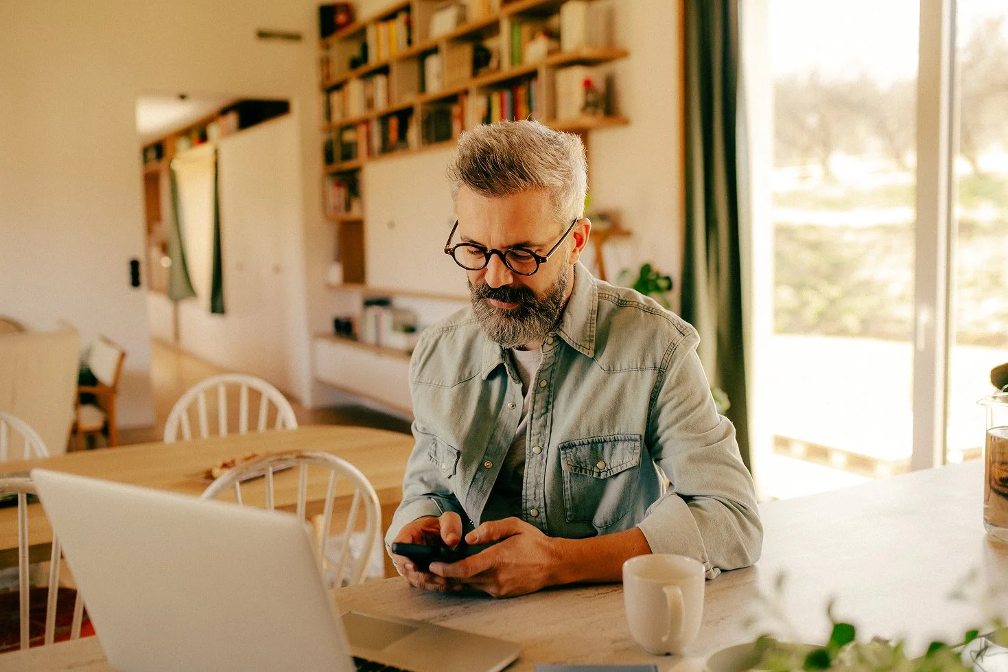 A middle-aged man with glasses and a gray beard sitting at a kitchen counter, looking at his phone, with a laptop, a white coffee mug, and a plant nearby. The background shows a bookshelf and a large window with green curtains letting in natural light.