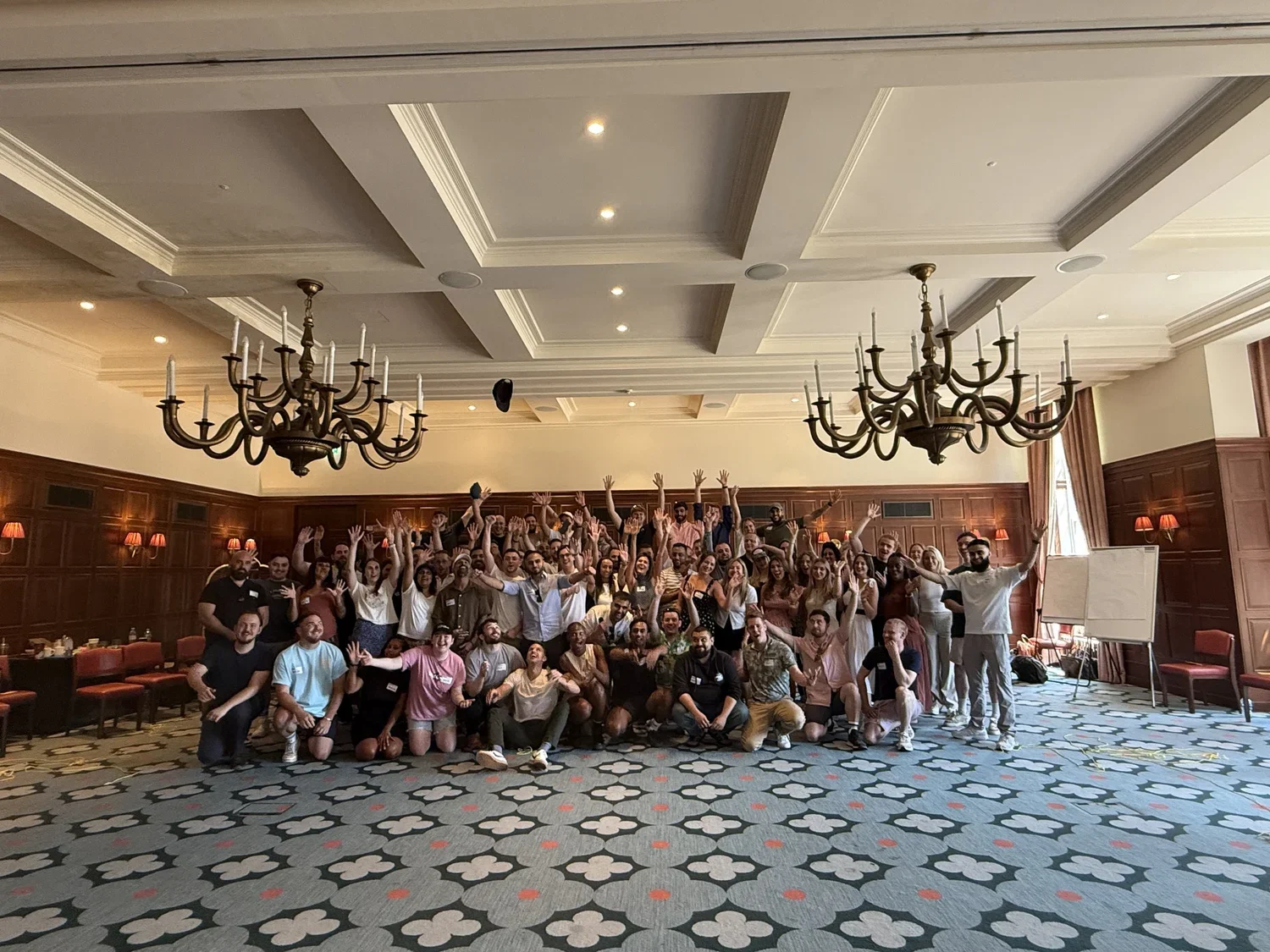 Large group of people gathered in a banquet hall, posing for a group photo with many raising their hands and smiling.