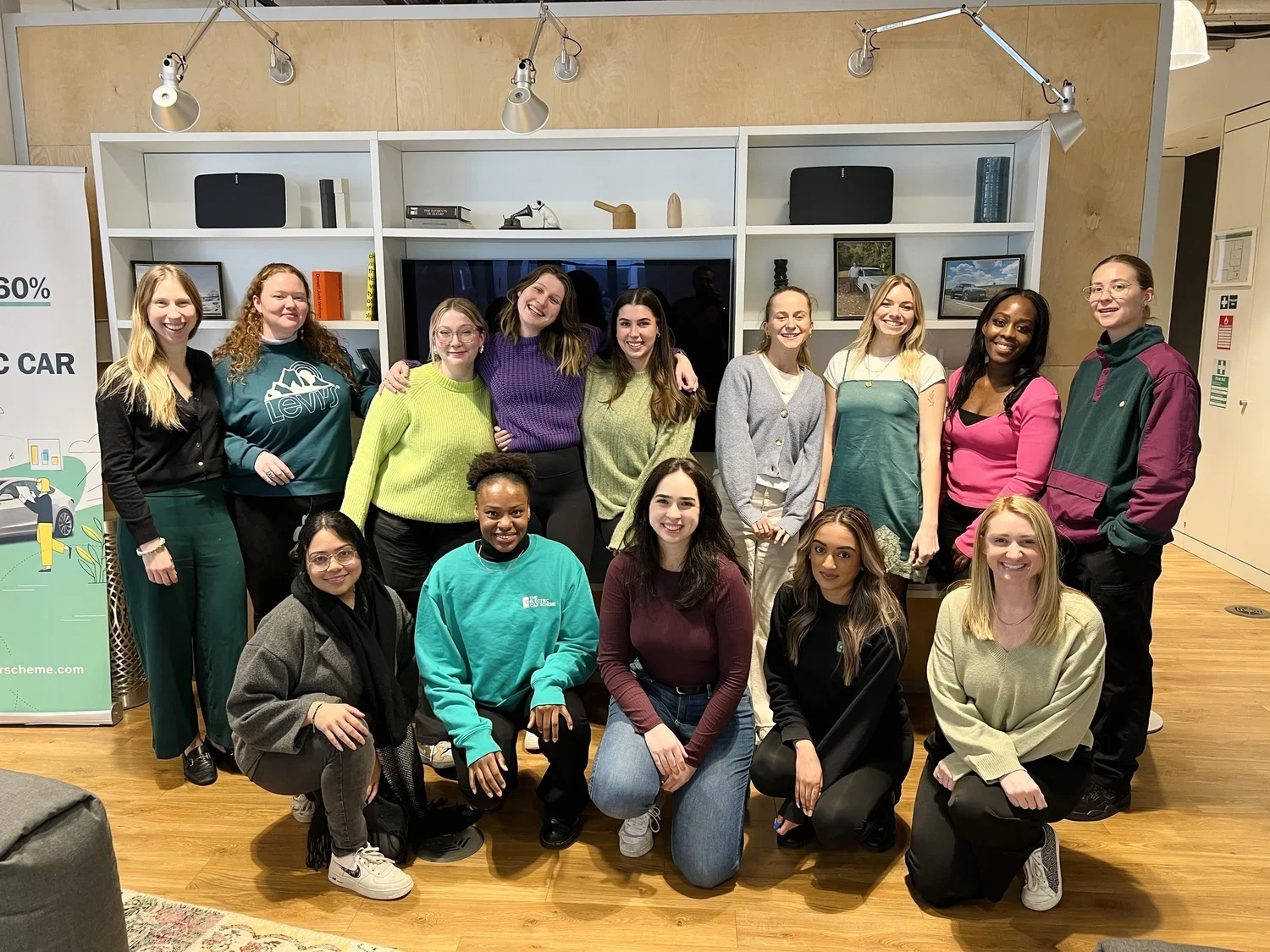 Group of 16 women smiling and posing together indoors in front of a white bookshelf and a television.