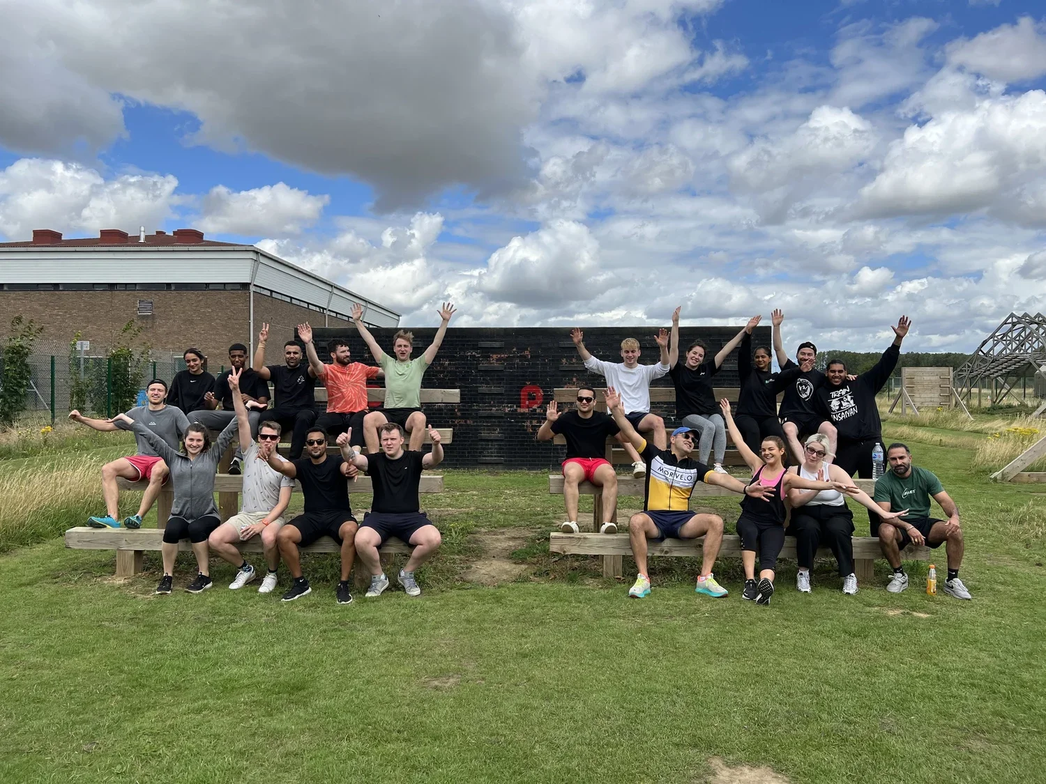 Group of 15 young adults posing outdoors in front of a brick wall monument, with arms raised, sitting and standing on wooden benches, under a partly cloudy sky.