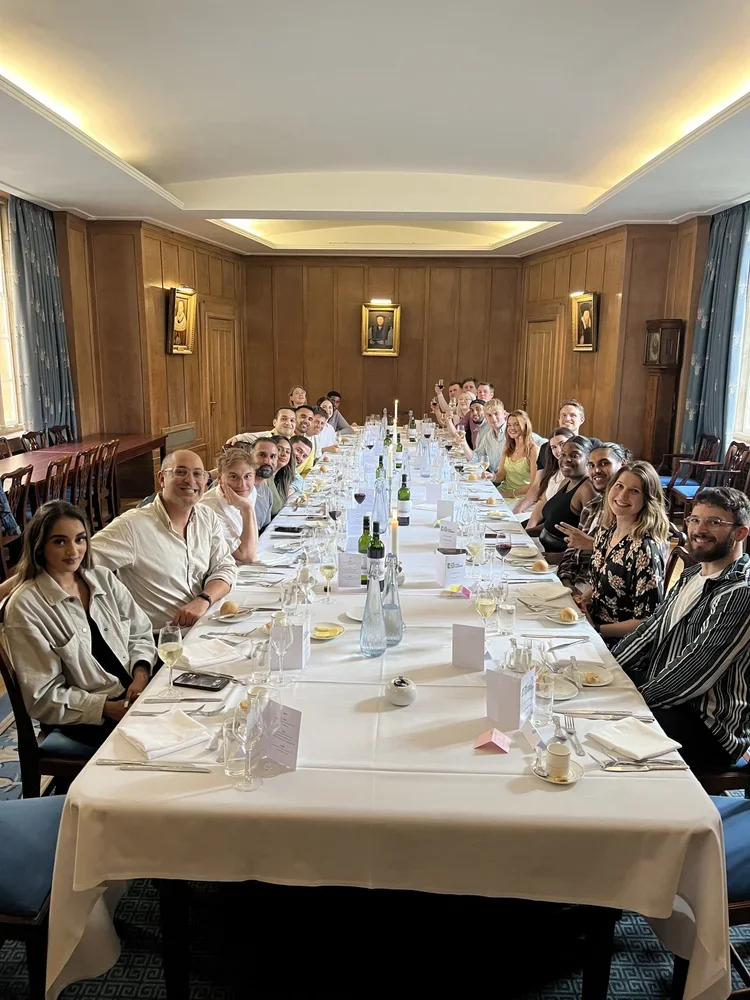 People sitting around a long dinner table in a wood-paneled room with portraits on the walls and large windows with curtains.