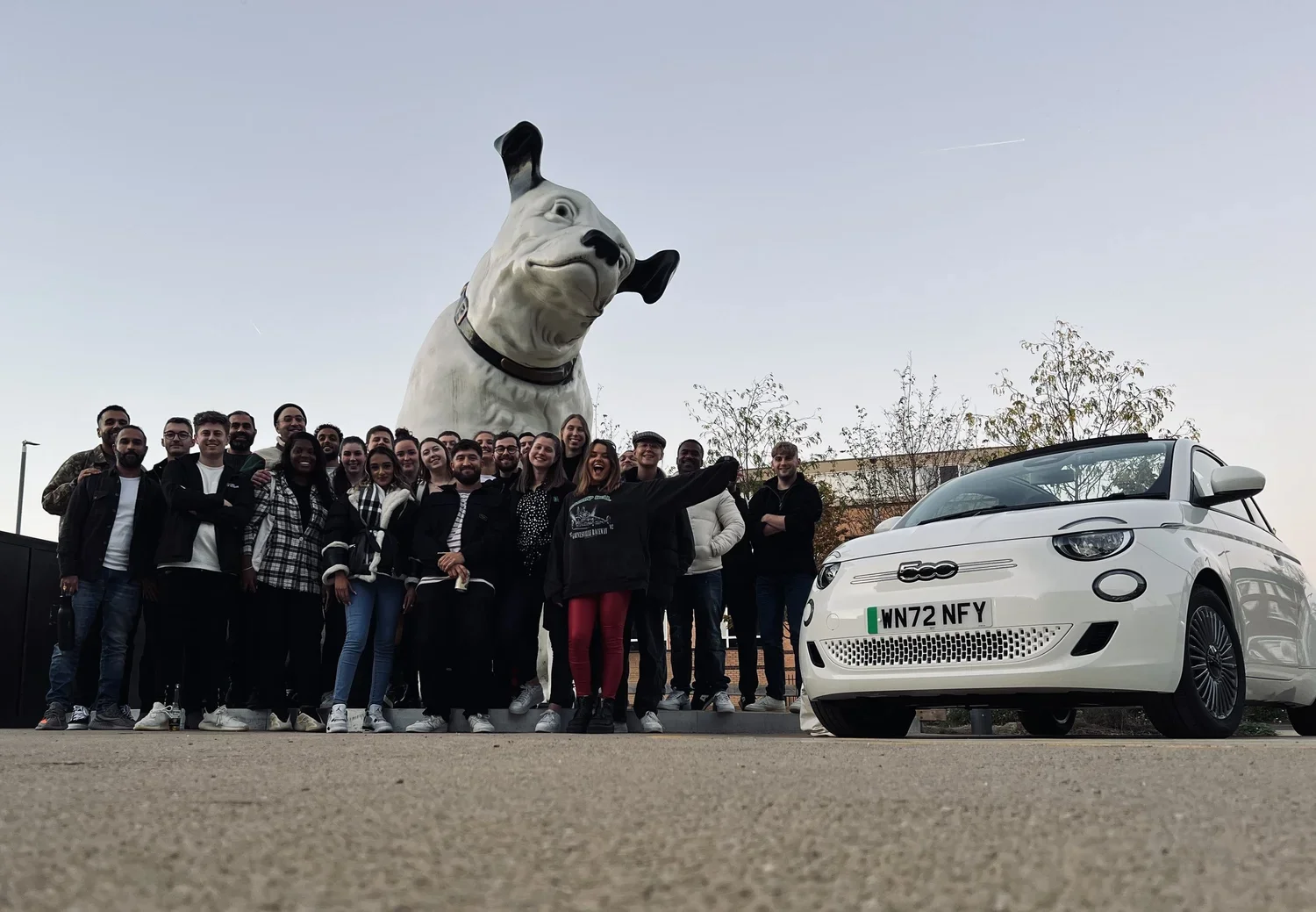 A group of people posing in front of a giant dog statue and a small white Fiat 500 car on a parking lot during daytime.