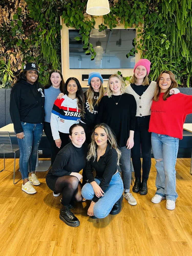 Group of nine women dressed casually, standing and sitting in a fitness or event space with green wall plants, a large screen, and wooden flooring.