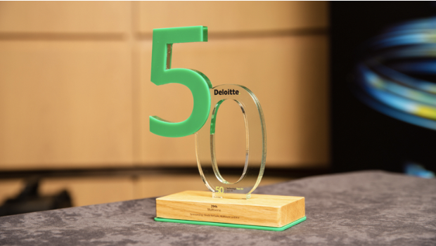 A transparent acrylic trophy with the number 50 and the words Deloitte, placed on a wooden base with a green front edge, on a gray surface.