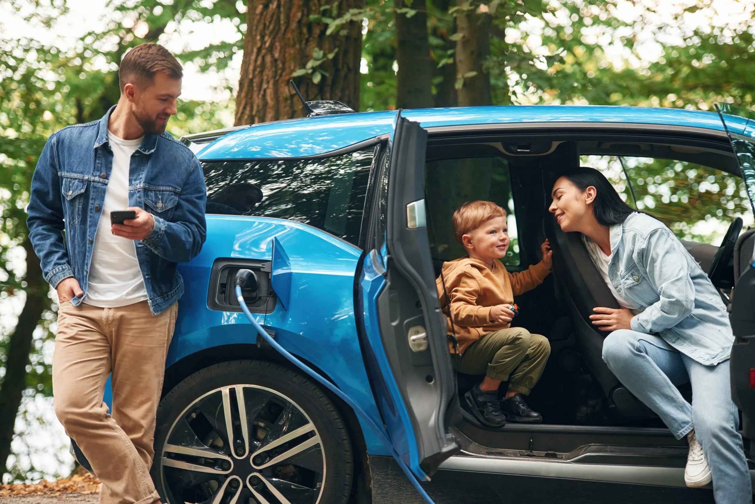 A family of three, with a young boy, and two adults, possibly parents, gathered near a blue electric car charging in a wooded area during daytime. The woman is inside the car talking to the boy, while the man is standing outside, holding a phone, smiling.
