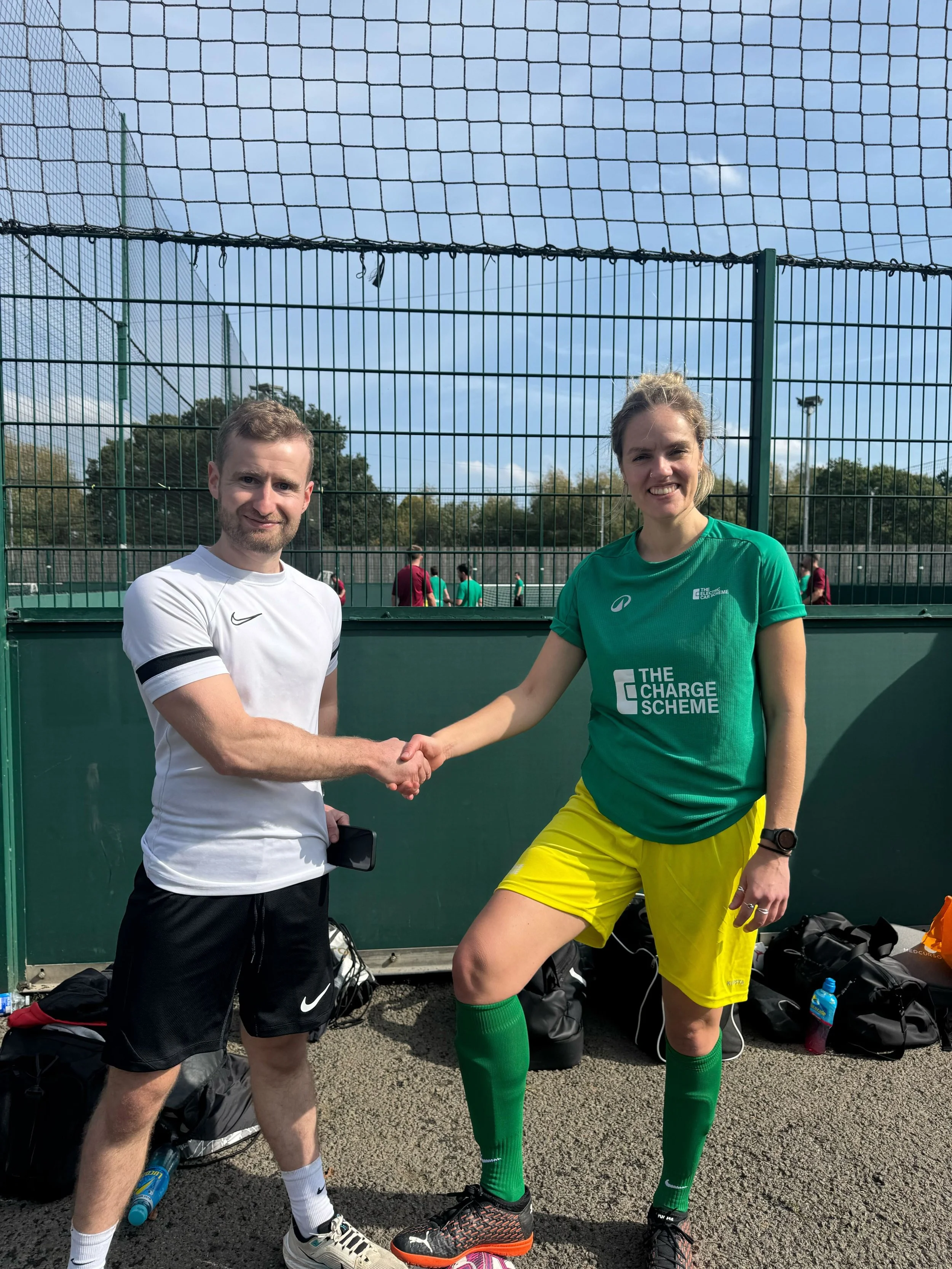Two people shaking hands on a tennis court, with sports bags and gear at their feet, and a net and trees in the background.