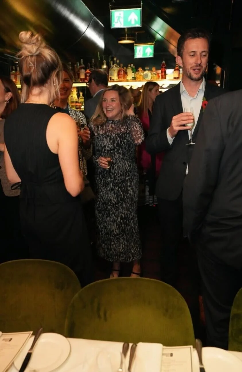 People gather at a bar or restaurant, socializing and celebrating, with drinks in hand and smiling guests dressed in formal attire. There are green exit signs hanging from the ceiling.