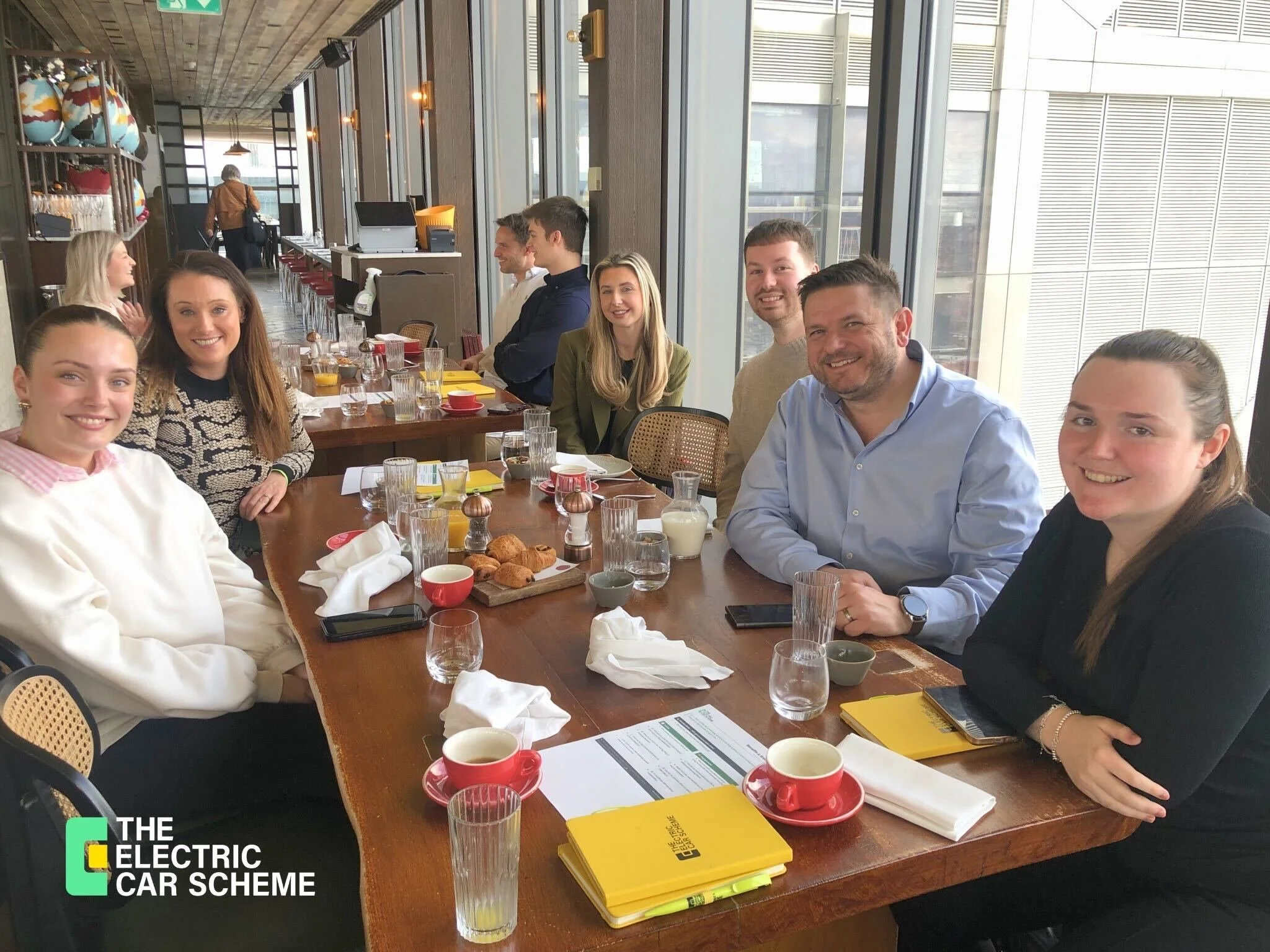 A group of people sitting at a long wooden table in a modern, bright cafe or restaurant with large windows. The table has plates with croissants, glasses, cups, napkins, and yellow booklets. There are people smiling and looking at the camera.