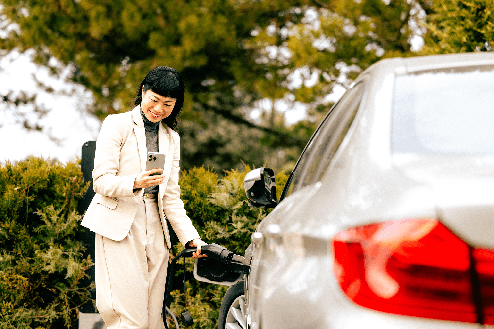 A woman with black hair and bangs, dressed in a beige blazer and gray turtleneck, is standing outdoors during fall, holding a smartphone in her right hand and plugging an electric car charger into a silver SUV. She is smiling and looking at her phone. The background features green trees and bushes with autumn colors.