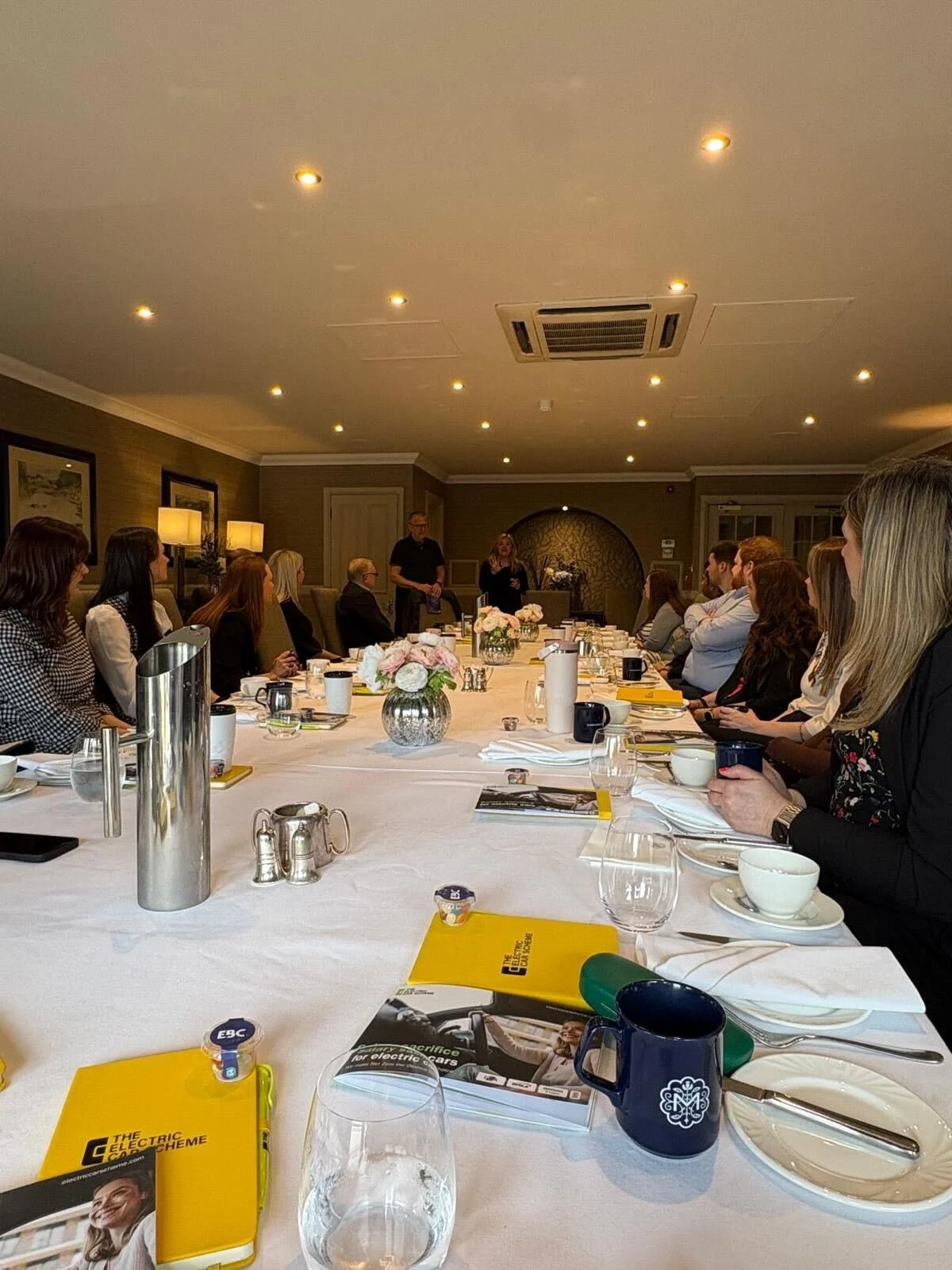 A business meeting or workshop taking place in a formal conference room with a long table. Attendees are seated on both sides, listening to a woman standing at the front. The table is set with cups, glasses, notebooks, and flowers. The room has warm 