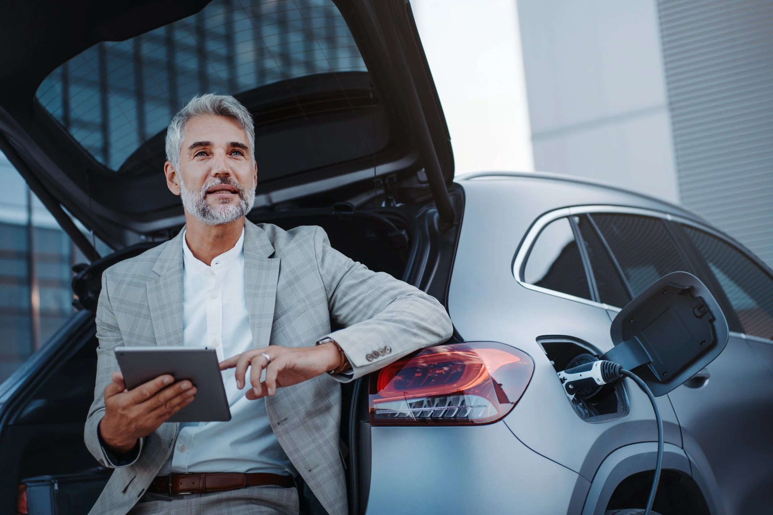 A middle-aged man with gray hair and a beard in a suit is standing next to an open electric car trunk, holding a tablet device, with a charging cable plugged into the car.